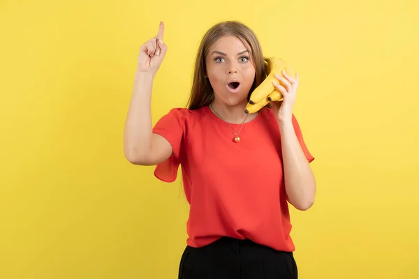 スマートフォンのように顔の近くにバナナを持っている若い女の子と黄色の背景の上にポーズ 高品質の写真 — ストック写真