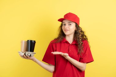Kırmızı tişörtlü ve şapkalı kıvırcık kız sarı duvarda kahve taşıyor. Yüksek kalite fotoğraf
