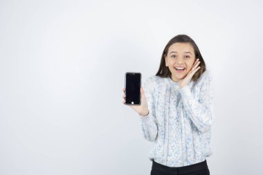 Süveter giyen kız beyaz arka planda akıllı telefonla poz veriyor.