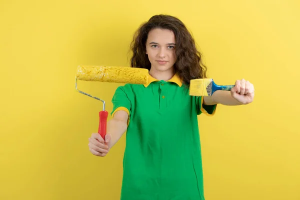 黄色の背景にペイントローラーとブラシを示す緑のTシャツの中の少女の肖像画 高品質の写真 — ストック写真