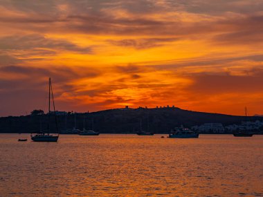 Yat ve tepelerin siluetleri deniz manzarası ve gökyüzünde turuncu bulutlar, batan güneş tarafından aydınlatılıyor..