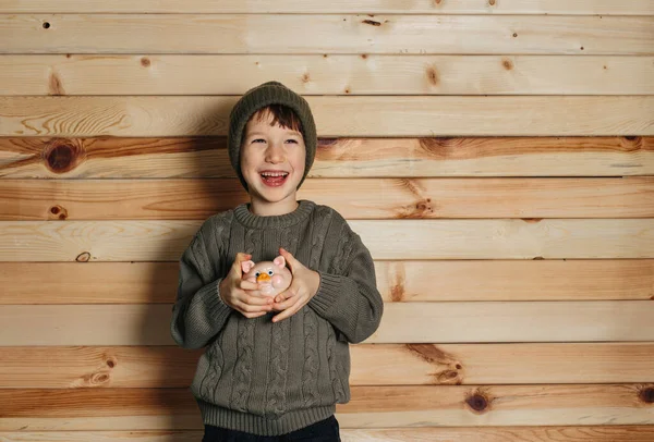 Портрет милого улыбающегося мальчика с копилкой на деревянном фоне. Ребенок в зеленой шляпе. — стоковое фото