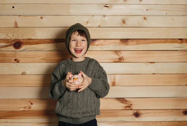 Retrato de menino sorridente bonito com banco porquinho no fundo de madeira. Criança com dinheiro em chapéu verde. Fotografias De Stock Royalty-Free