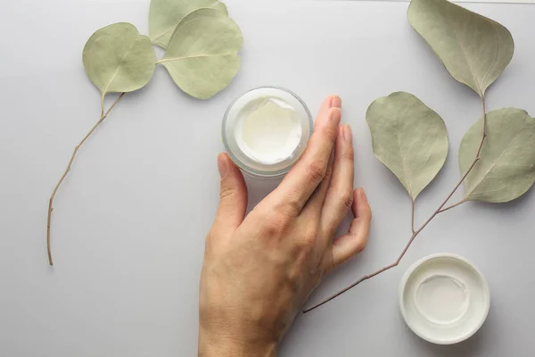 Vista dall'alto di un barattolo di crema su sfondo bianco, con foglie di eucalipto e mano femminile. — Foto Stock