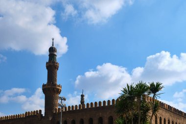 Geleneksel İslami mimarinin üzerinde yükselen tarihi cami minaresinin alçak açılı görüntüsü, parlak mavi gökyüzünün altında, dağınık bulutlarla çevrili palmiye ağaçları tarafından çerçevelenmiş.