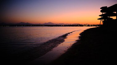 Gün batımında sahilde sakin dalgalar sahile vuruyor. Sıcak turuncu ve mor renkler gökyüzünü doldurarak uzak kara parçalarının siluetleriyle sakin bir kıyı atmosferi yaratıyor.