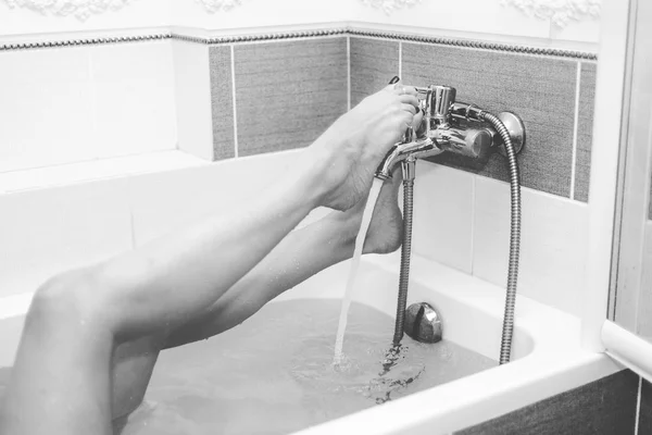 Monochrome image. Shapely female legs with pedicure over the bath in the water, switch on the mixer ta