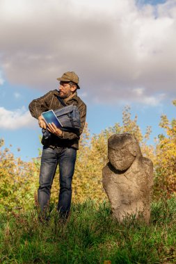 Kareli ceketli sakallı turist bulutlu bir gökyüzüne karşı yeşil bir yamaçta İskitler döneminden kalma taş bir heykelin yanında duruyor.