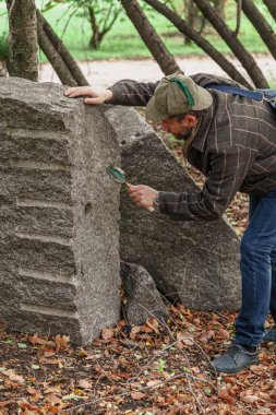 Jeolog, büyük bir gri kaya parçasını kemik sapındaki büyük bir büyüteçle inceliyor. Delik izleri ston üzerinde görülebilir.