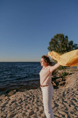 Geniş bir nehrin kumlu sahilinde uzun kızıl saçlı, parlak sarı ceketli, iri yarı, güzel bir kızın portresi.