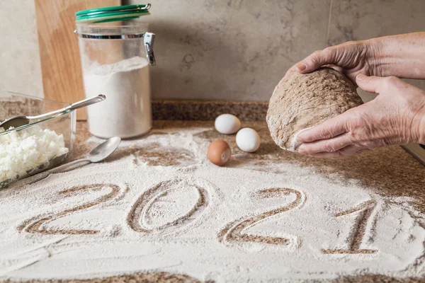 Mutfakta yeni yıl. 2021 un ile yazılmıştır. Elleri hamuru tutuyor. Granit tezgahta duran yumurta ve cam kase.