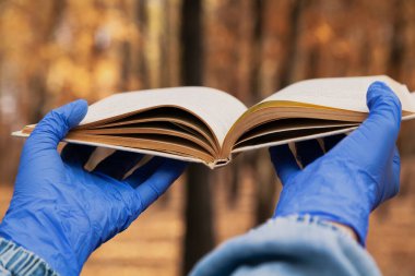 Tecritteyken okuyorum. Mavi kauçuk eldivenli eller sonbahar ormanında açık bir kitap tutuyor. Yakın plan.