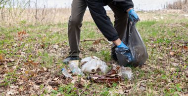 Doğayı çöpten kurtarıyoruz. Mavi lastik eldivenli bir adam, bir kuponun kıyısında siyah bir çantada plastik çöp topluyor.
