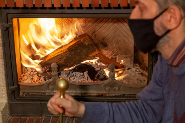 We are sitting at home on self-isolation. A man in a black protective mask looks at the fire in the burning fireplace. Holds a poker in his hand