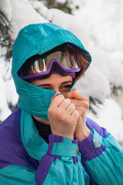 Uzun bir çam ağacının altında kapüşon takan kayak tulumlu güzel bir genç kadın. Çam dalında kar var.