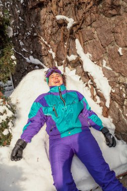 Karda dinlen. Tulum giymiş güzel bir kız sırtı karda yatıyor ve gülümsüyor.