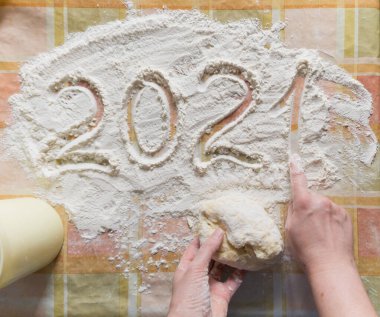 Mutfakta yeni yıl. El, damalı masa örtüsünün üzerine 2021 yazıyor. Sol elinde bir parça hamur var. Fotoğraf smartphon ile çekildi