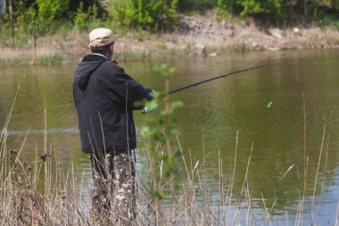 Ceketli bir adam, sonbaharda bir su deposunun kıyısında dönen bir çubukla balık tutuyor.