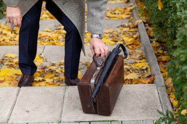 Antik deri valiz sonbahar parkının merdiveninde duruyor. Üstte bir şemsiye bastonu var. Açık renk ceketli bir beyefendi ona eliyle dokunur.