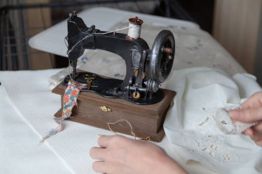 Beyaz iplik makarası olan klasik dikiş makinesi beyaz bir kumaşın üzerinde duruyor. Dişinin elinde pıhtı var.