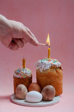 Pembe arka planda iki Paskalya pastası ve yumurta standı. Mumlar Paskalya pastalarının içine konur. Bir el kibritle bir mum yakar. Ön planda beyaz çiçekli bir ağaç dalı var.