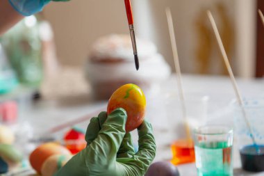 Kauçuk mavi eldivenli eller bir yumurta ve boya fırçası tutuyor. Ev yapımı Paskalya yumurtası ve renkli likörlü bardaklar.