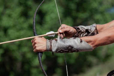 Yumuşak zırhla korunan güçlü erkek elleri bulanık yeşil bir sırt grubuna uzanan antika bir yay telini gerer.