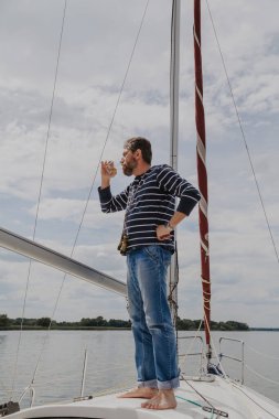Çizgili kazaklı sakallı bir iş adamı beyaz bir yatın burnunda duruyor ve camdan viski içiyor.