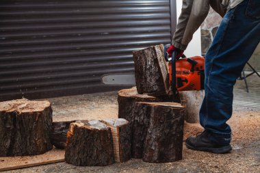 Elektrikli testere çalışıyor. Bir adam ağaç gövdesinin bir parçasını elektrikli testereyle kesiyor. Parça avludaki büyük bir ağaç kütüğünün üzerinde duruyor.