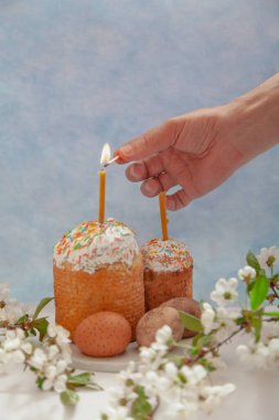 Mavi arka planda iki Paskalya pastası ve yumurta standı. Mumlar Paskalya pastalarının içine konur. Bir el kibritle bir mum yakar. Ön planda beyaz çiçekli bir ağaç dalı var.