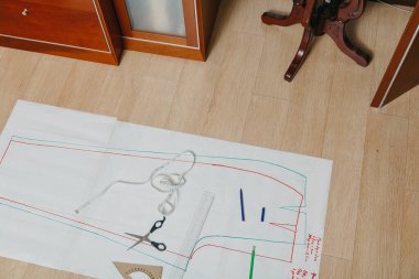 Dikiş izleri yerde yatıyor. Kağıt bir çarşafa bir giysi silueti çizilir. Üstte cetvel, makas ve esnek bir santimet var.