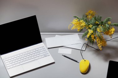 Sarı fareli laptop gri bir masada duruyor. Yakınlarda bir cep telefonu kartı ve bir buket sarı mimoza var. Yukarıdan Görünüm