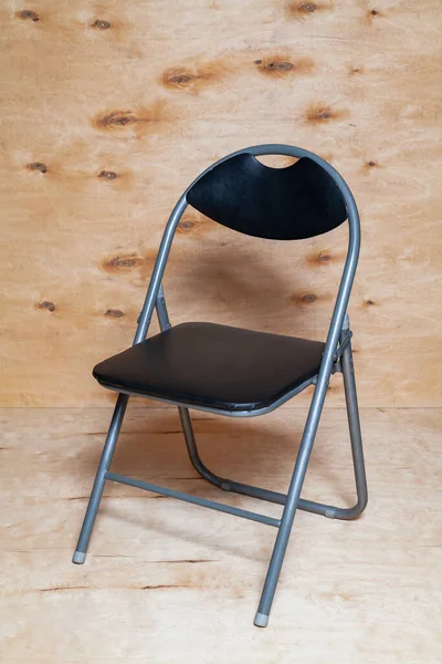 Art Nouveau folding chair stands against a background of light plywood. The chair has an arched back and a black sea