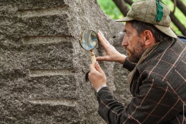 Jeolog, büyük bir gri kaya parçasını kemik sapındaki büyük bir büyüteçle inceliyor. Delik izleri ston üzerinde görülebilir.