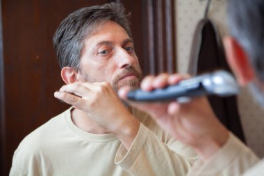 Tıraş makinesiyle sakalını kesiyorum. Bej tişörtlü hoş bir beyefendi, banyoda büyük bir aynanın önünde daktilo kullanarak gri sakalını tek başına kesiyor.