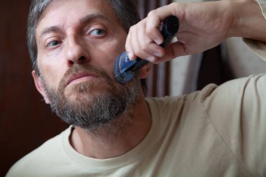 Saç kesim sakal kesme makinesi. Gri sakalını tek başına kesen bir adam hortumu kapalı bir makine kullanıyor.