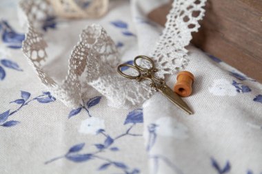 İnce dantel örgü ve sarı vintage makas mavi çiçek desenli beyaz kumaşın üzerinde yatar. Yakınlarda ahşap bir kutu ve kakao var.