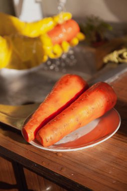 İki büyük kırmızı havuç bir tabakta yakın planda duruyor. Arka planda eldiven giymiş bir elin bulanık görüntüsü var.