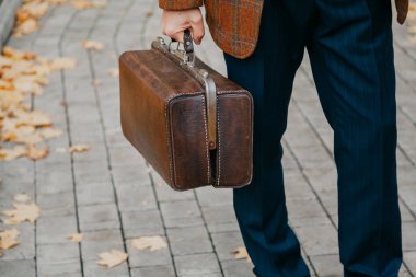 Şık bir ceketi olan beyefendi sonbahar öğleden sonra meydanın ortasında duruyor. Elinde kaliteli bir halı çantası tutuyordu. Kapat.