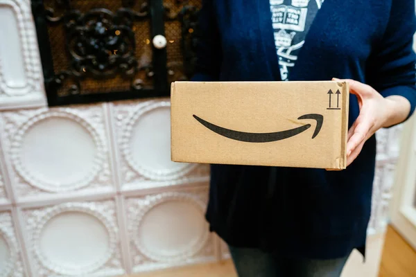 Seitenansicht einer eleganten Frau, die ein Amazon Prime-Paket mit einem pfeilgrinsenden Schriftzug und einem Luxus-Ofen im Hintergrund als Geschenk hält — Stockfoto