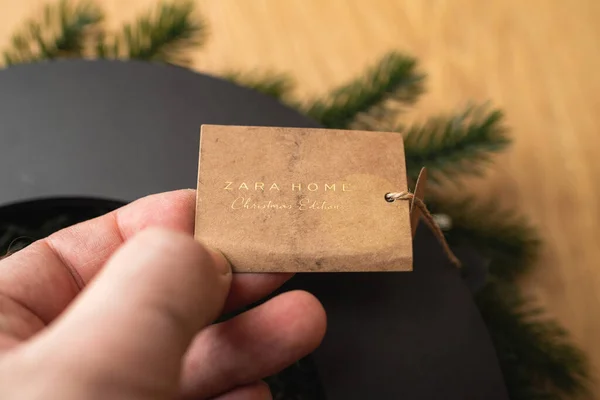 POV männliche Hand hält Zara home Christmas Edition Preisschild auf dem Recyclingpapier auf einem dekorativen Kranz für zu Hause — Stockfoto