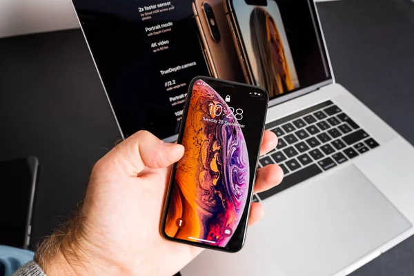 POV mužská ruka drží nejnovější Apple Computers iPhone Max smartphone s běžícím MacBook Pro v pozadí velký displej sítnice OLED — Stock fotografie