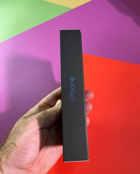 Мужчина держит руку на красно-фиолетовом желтом фоне нового смартфона Apple iPhone 12 Pro в новой упаковке - тонкая сторона нового экологического пакета — стоковое фото