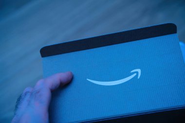 POV erkek, karton panelde Amazon Şirketi amblemi olan karton bir kutu tutuyor - mavi arkaplan