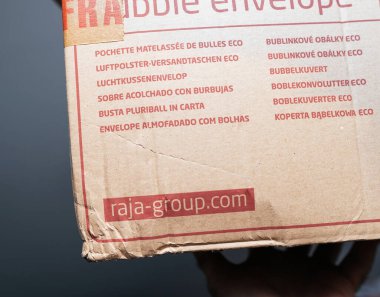 Paris, Fransa - 13 Aralık 2024: Çok dilli eko baloncuklu zarf metnini ve tarafsız bir arkaplana karşı elle tutulan görünür Raja Grubu web sitesinin logosunu gösteren karton bir nakliye kutusunun yakın çekimi.