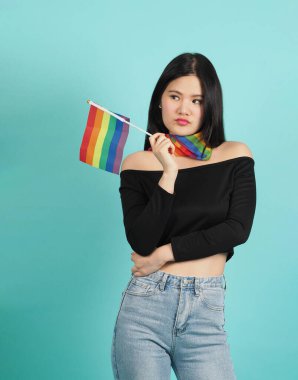 LGBT bayrağı tutan genç bir kızın portresi mavi yeşil arka plan stüdyosunun karşısında duruyor. Boynunda gökkuşağı eşarbı olan Asyalı LGBTQ kadını. Zeki, neşeli ve enerjik görün. LGBTQ kavramı.