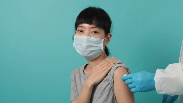 Vaccine Concept. Woman waiting for coronavirus vaccine injection by doctor. Asian woman with medical mask open shoulder and upper arm on blue green background. Waiting for vaccination.