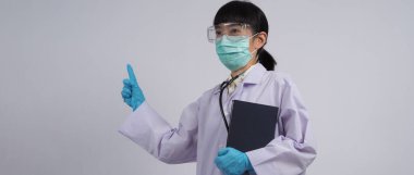 Üniformalı doktor boş bir bölgeyi işaret ediyor. Salgın virüsünü önlemek için mavi lateks eldivenler, temiz gözlükler ve yeşil N95 maskesi takıyor. Asyalı doktor. El hareketi. Tıbbi steteskop. Virüs Kitabı