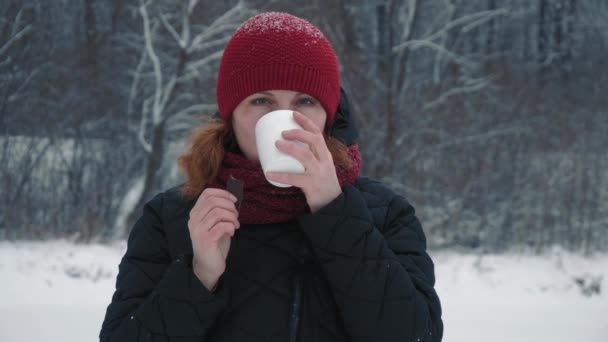 Vrouw met een rode warme hoed drinkt warme thee of koffie uit een kopje in een besneeuwd bos, wil zich in de winter buiten opwarmen met een mok warme drank. Kerstvakantie. Langzame beweging — Stockvideo