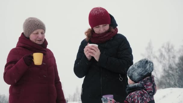 Famiglia felice bere tè caldo all'aperto in inverno. Di attraente signora anziana con la figlia e il nipote bere tè caldo o caffè da una tazza in una foresta innevata — Video Stock